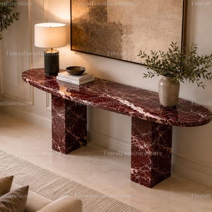 May include: A long, oval-shaped console table crafted from deep red marble with prominent white veining. The table features two thick, rectangular pedestal bases and a smooth, polished surface supporting a lamp, books, and a small bowl.