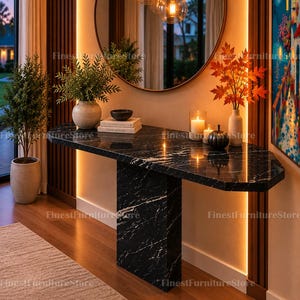 May include: A black marble console table with white veining features a rectangular top and a central pedestal base. It is decorated with a potted green plant, stacked books, a small bowl, two candles, and a vase with orange autumn leaves.