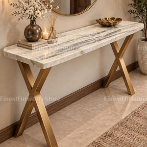 May include: A console table featuring a rectangular white marble top with grey veining and two gold-toned metal legs arranged in an X-shape. The table surface holds a dark vase with white flowers, a small candle, and a decorative gold bowl.