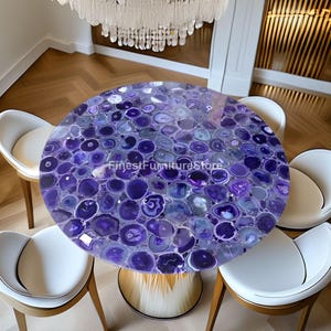 May include: A round dining table with a purple agate stone top, supported by a gold and cream-colored base. The table is surrounded by four white chairs with wooden legs. A crystal chandelier hangs above the table.