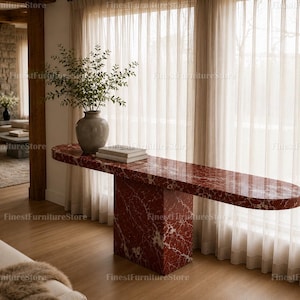 May include: A long, narrow console table crafted from deep red marble with prominent white veining. The table features a thick, rectangular pedestal base supporting a smooth, elongated oval top. A ceramic vase with green foliage sits on top.