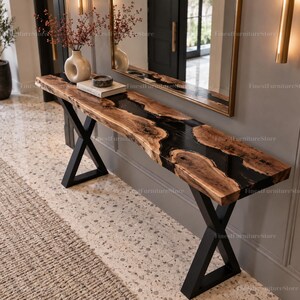 Puede incluir: Una consola larga con una superficie de madera natural de borde vivo y una incrustación central de resina negra. La mesa está sostenida por dos patas de metal negro en forma de X. La veta de la madera es visible, mostrando su textura orgánica.