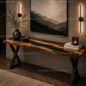 Pode incluir: Uma consola comprida com um tampo de madeira natural e uma incrustação central de resina verde-escura. A mesa é suportada por duas pernas de metal preto em forma de X. Em cima, encontram-se um vaso escuro com vegetação, uma taça e dois livros.