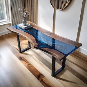 Può includere: Un tavolo in legno fatto a mano con un design a fiume in resina blu. Il tavolo presenta un bordo naturale, un bordo in legno scuro e gambe in metallo nero. Un vaso con fiori e un libro sono sul tavolo.