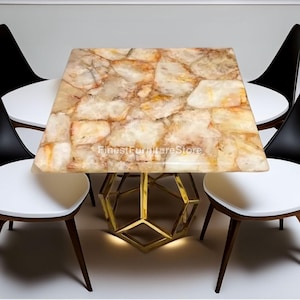 May include: A square table with a natural stone top in shades of beige, cream, and brown. The table has a geometric gold-colored metal base. Four modern chairs with white seats and black backs surround the table.