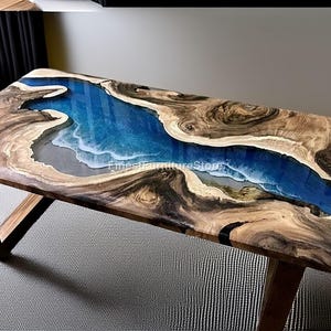 Op de afbeelding: Rechthoekige houten salontafel met een blauw epoxyhars rivierontwerp. De tafel heeft een natuurlijke houtnerf rand en schuine houten poten. Het hars rivierontwerp lijkt op een strandscène met witte golven en een blauwe oceaan.