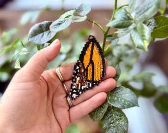 May include: A beaded earring in the shape of a monarch butterfly wing. The earring features orange, black, white, and brown beads, with a gold-colored hook. The earring is held in front of a leafy green plant.