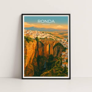 Puede incluir: Una impresión artística enmarcada que presenta una vista panorámica de Ronda, Málaga. La obra de arte representa un puente que cruza un profundo desfiladero, con edificios y acantilados en tonos cálidos de naranja, amarillo y blanco. El cielo transita de azul a naranja. El texto en la parte superior dice "RONDA MALAGA."
