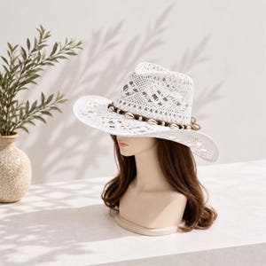 May include: A white crocheted fedora hat featuring an openwork pattern throughout the crown and brim. The hat is accented with a decorative band composed of small brown wooden beads and light-colored cowrie shells encircling the base.