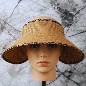 May include: A straw visor with a wide brim and a leopard print trim. The visor is tan and has a curved shape, designed to shield the face from the sun. The leopard print adds a stylish accent.