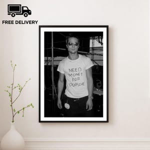 May include: Black and white framed print featuring a person wearing sunglasses and a white t-shirt with the handwritten text "NEED MONEY FOR PORSCHE". The image is a portrait, with a black frame and a white background.