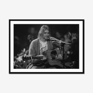 Peut inclure: Une photographie encadrée en noir et blanc montrant un musicien aux cheveux mi-longs portant un cardigan sur un t-shirt, jouant de la guitare acoustique derrière un pied de micro. La scène est capturée dans un environnement peu éclairé.