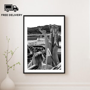 May include: Monochrome framed print showing a woman in a convertible with her legs raised, wearing cowboy boots. The image is a photograph of a vintage car, evoking a sense of freedom.