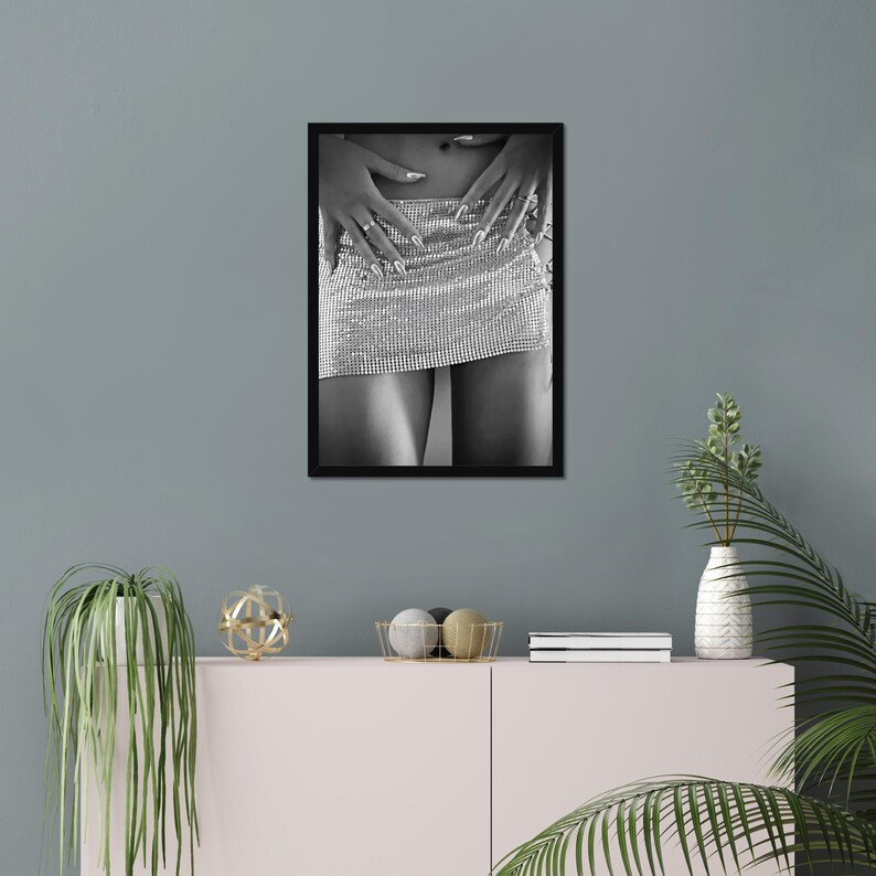 May include: Black and white framed photograph of a silver rhinestone mini skirt. The skirt is the focal point, with hands gently resting on it. The image is a close-up, emphasizing the texture and sparkle of the skirt.