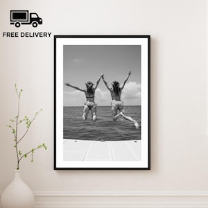 May include: Monochrome framed print showing two people jumping into the sea, holding hands. The image is set against a bright sky and water. The frame is black and the print is displayed on a white wall.