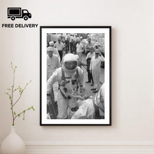 Puede incluir: Impresión enmarcada en blanco y negro de un astronauta con un traje espacial, rodeado de personas con uniformes blancos. El astronauta, con un casco y traje detallados, es el foco. La imagen es una fotografía vintage, probablemente de la era espacial.