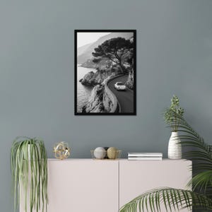 May include: Black and white framed print of a coastal road winding along a cliffside. A white car travels on the road, with a large tree and the sea visible. The print is displayed on a gray wall.