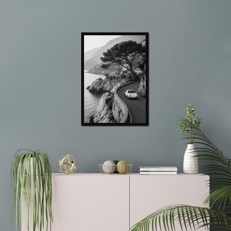 May include: Black and white framed print of a coastal road winding along a cliffside. A white car travels on the road, with a large tree and the sea visible. The print is displayed on a gray wall.