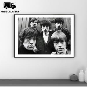 Puede incluir: Fotografía en blanco y negro enmarcada que presenta a cinco hombres. Los hombres visten ropa oscura y tienen el pelo largo. La fotografía se exhibe en una pared blanca sobre una estantería gris.