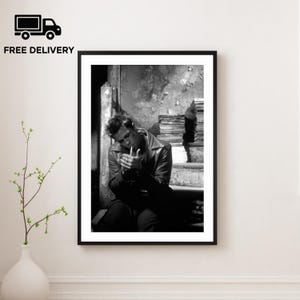 Könnte beinhalten: Schwarz-Weiß-Druck in einem Rahmen, der einen Mann beim Anzünden einer Zigarette zeigt. Der Mann, in einer Lederjacke, sitzt auf einer Treppe. Das Bild hat eine Vintage-Film-Noir-Ästhetik. Die Worte "FREE DELIVERY" befinden sich in der oberen linken Ecke.