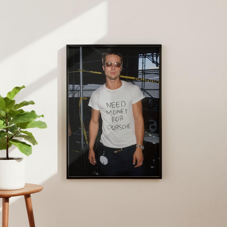 May include: Framed photograph of a person wearing sunglasses and a white t-shirt with the handwritten text "NEED MONEY FOR PORSCHE". The person is standing in front of a construction site.