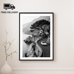 May include: Black and white framed print of a coastal road with a white car driving along the winding path. A large tree and rocky cliffs border the road, with the sea visible on the left. The image is in a black frame.