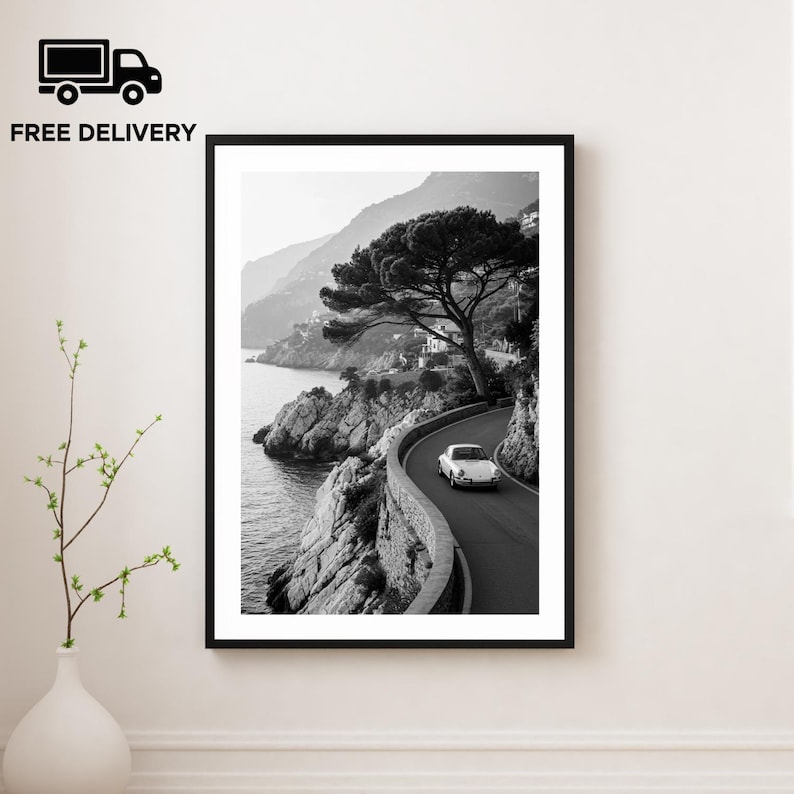 May include: Black and white framed print of a coastal road with a white car driving along the winding path. A large tree and rocky cliffs border the road, with the sea visible on the left. The image is in a black frame.