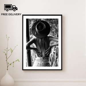 May include: Monochrome framed print showing a person with long blonde hair, wearing a hat, and denim jeans. The person's back is to the viewer, hands on the lower back. The background is a blurred outdoor scene.