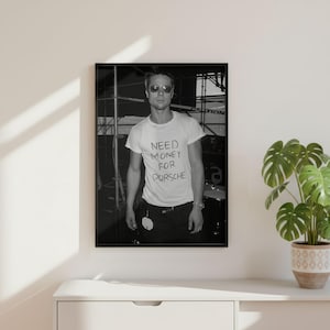 May include: Black and white framed print featuring a person wearing sunglasses and a white t-shirt with the handwritten text "Need Money For Porsche". The image is a portrait, with the person standing in front of a blurred background.
