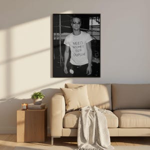 May include: Black and white framed print of a person wearing sunglasses and a white t-shirt with the text "NEED MONEY FOR PORSCHE". The print is hanging on a wall above a beige sofa with pillows and a blanket.