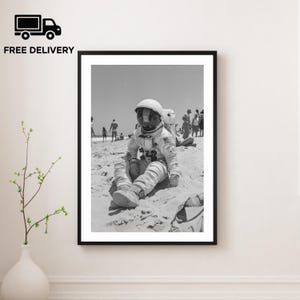 Puede incluir: Impresión enmarcada en blanco y negro de un astronauta sentado en una playa arenosa. El astronauta lleva un traje espacial y un casco. Hay gente en el fondo. La impresión se muestra en una pared blanca.