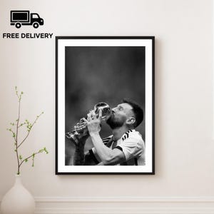 Może przedstawiać: Czarno-biała oprawiona grafika przedstawiająca piłkarza całującego trofeum. Obraz jest zbliżeniem zawodnika, z trofeum w centrum uwagi. Słowa "FREE DELIVERY" znajdują się w lewym górnym rogu.
