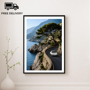 May include: Framed print of a coastal road with a white car driving along the winding path. The image features a scenic view of the ocean, mountains, and a large tree. The print is in a black frame and is displayed on a white wall.