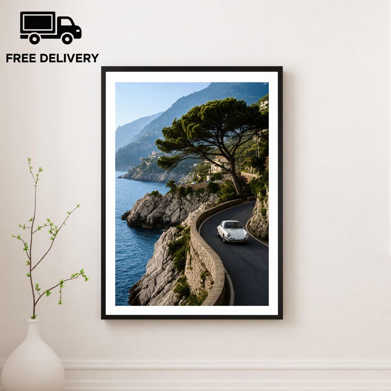 May include: Framed print of a coastal road with a white car driving along the winding path. The image features a scenic view of the ocean, mountains, and a large tree. The print is in a black frame and is displayed on a white wall.