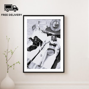 Puede incluir: Impresión enmarcada en blanco y negro que muestra un chimpancé y una mujer con gafas de sol relajándose en tumbonas. La mujer lleva un bikini. Un salvavidas y sillas de playa están en el fondo. La impresión se muestra en una pared blanca.