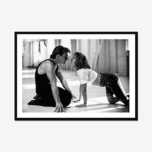 May include: Monochrome framed print depicting a man and a woman in a dance pose. The man kneels, dressed in a black vest and trousers. The woman is bent over, wearing a white top and jeans. The image is a still from a film.