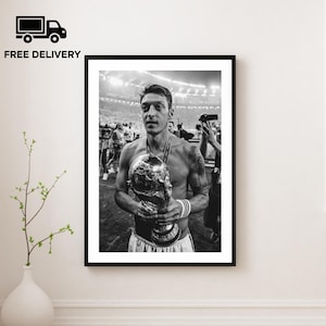 May include: Black and white framed print of a soccer player holding the World Cup trophy. The player is shirtless, wearing a medal, and surrounded by people. The image is in a stadium setting.