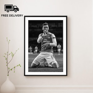 May include: Black and white framed print of a soccer player kneeling on the field, celebrating a goal. The player wears a red and white jersey with the Emirates logo. The image is displayed on a white wall.