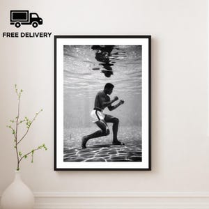 Könnte beinhalten: Schwarz-Weiß-Druck eines Boxers unter Wasser, in Kampfhaltung. Der Boxer trägt weiße Shorts mit schwarzem Streifen. Das Bild ist vor einem Hintergrund aus welligem Wasser angeordnet.