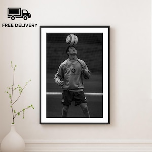 Może przedstawiać: Czarno-biała oprawiona grafika przedstawiająca piłkarza balansującego piłką nożną na głowie. Zawodnik ma na sobie koszulkę z długim rękawem i szorty. Grafika jest wyświetlana na białej ścianie. Słowa "FREE DELIVERY" znajdują się w lewym górnym rogu.