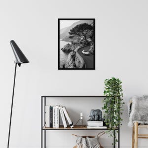 May include: Black and white framed print of a coastal road with a vintage car. The image captures a winding road along a rocky coastline, with a classic car driving on the road. A large tree is on the right side of the road.