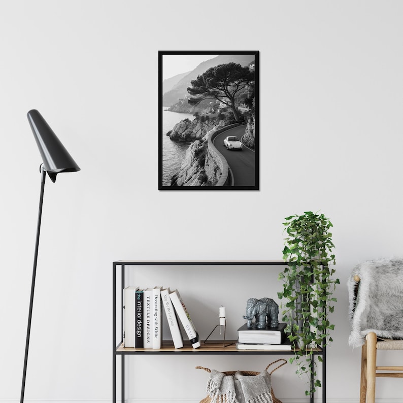 May include: Black and white framed print of a coastal road with a vintage car. The image captures a winding road along a rocky coastline, with a classic car driving on the road. A large tree is on the right side of the road.