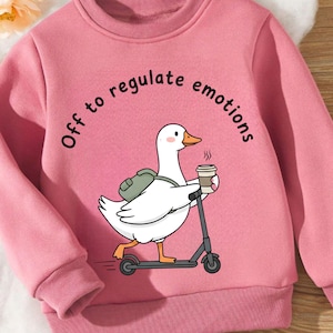 May include: Pink sweatshirt with the phrase "Off to regulate emotions." A white goose wearing a backpack and holding a coffee cup is riding a scooter. The sweatshirt has a crew neck and long sleeves.