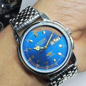 May include: A Citizen automatic wristwatch with a blue dial and a silver-tone stainless steel band. The watch face displays the day and date, with gold-tone hands and markers. The watch is marked "21 Jewels" and "Japan".