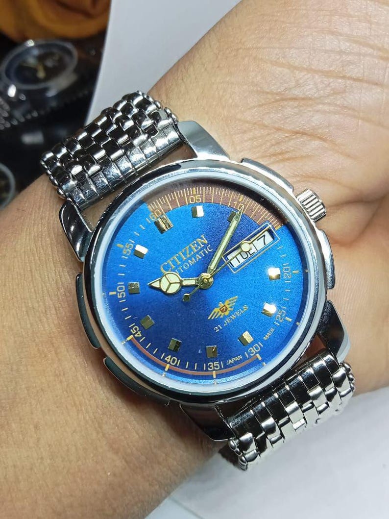 May include: A Citizen automatic wristwatch with a blue dial and a silver-tone stainless steel band. The watch face displays the day and date, with gold-tone hands and markers. The watch is marked "21 Jewels" and "Japan".