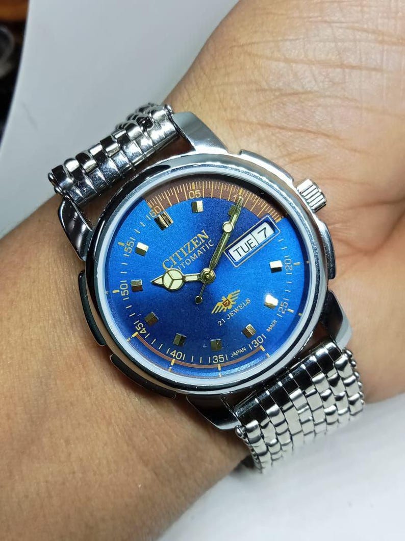 May include: A Citizen automatic wristwatch with a blue dial and silver-tone metal band. The watch face displays the day of the week and the time. The watch has a silver-tone case and a textured metal band.