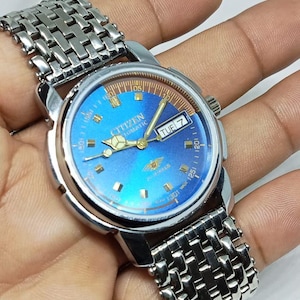May include: A vintage Citizen automatic watch with a blue dial and silver-tone stainless steel band. The watch face displays the day of the week and features gold-tone hands and markers. The watch is a classic timepiece.