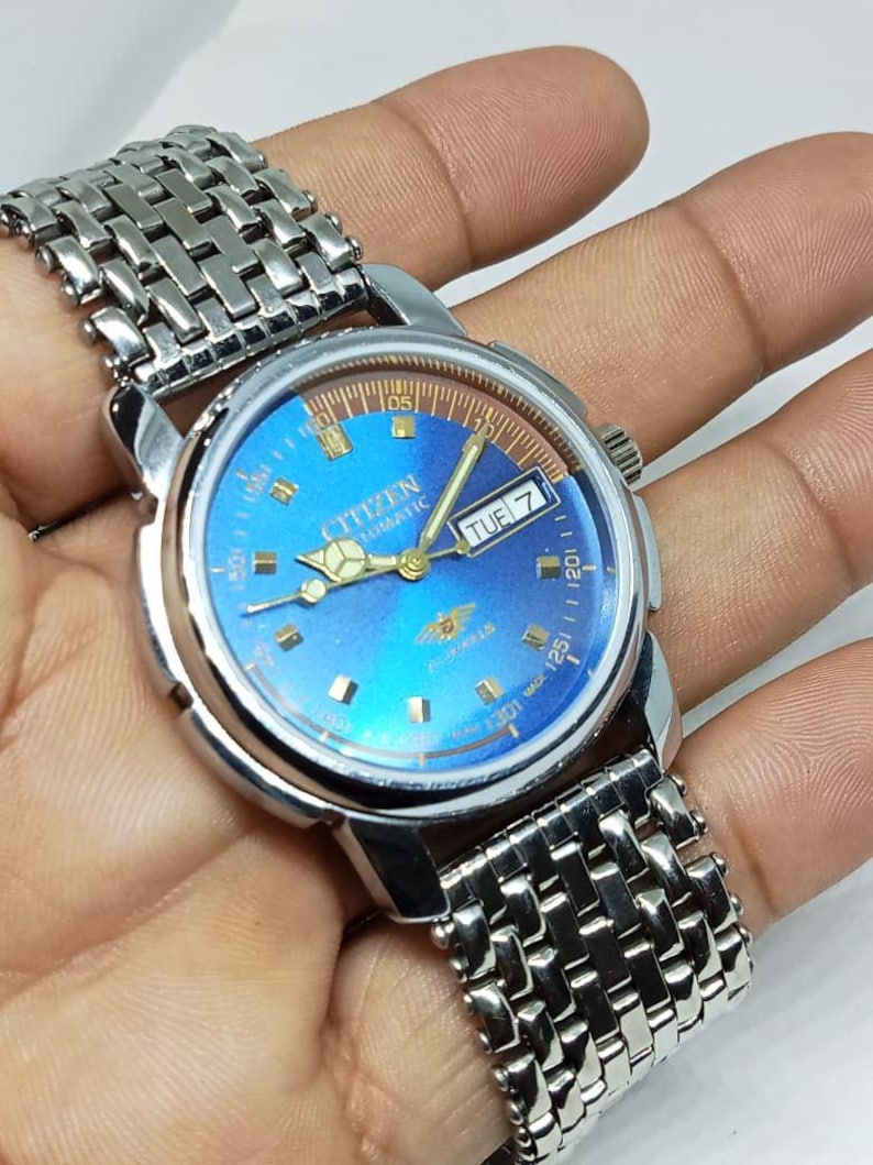 May include: A vintage Citizen automatic watch with a blue dial and silver-tone stainless steel band. The watch face displays the day of the week and features gold-tone hands and markers. The watch is a classic timepiece.