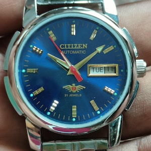 May include: A Citizen automatic wristwatch with a blue dial and silver-tone stainless steel band. The watch face displays the day and date, with gold-tone hands and markers. The watch has the text "Citizen Automatic" and "21 Jewels" on the dial.