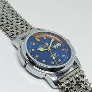 Op de afbeelding: Een vintage Citizen automatisch horloge met een blauwe wijzerplaat en een zilverkleurige kast en band. De wijzerplaat toont de dag van de week en heeft goudkleurige wijzers en markeringen. De band is een geweven metalen mesh.